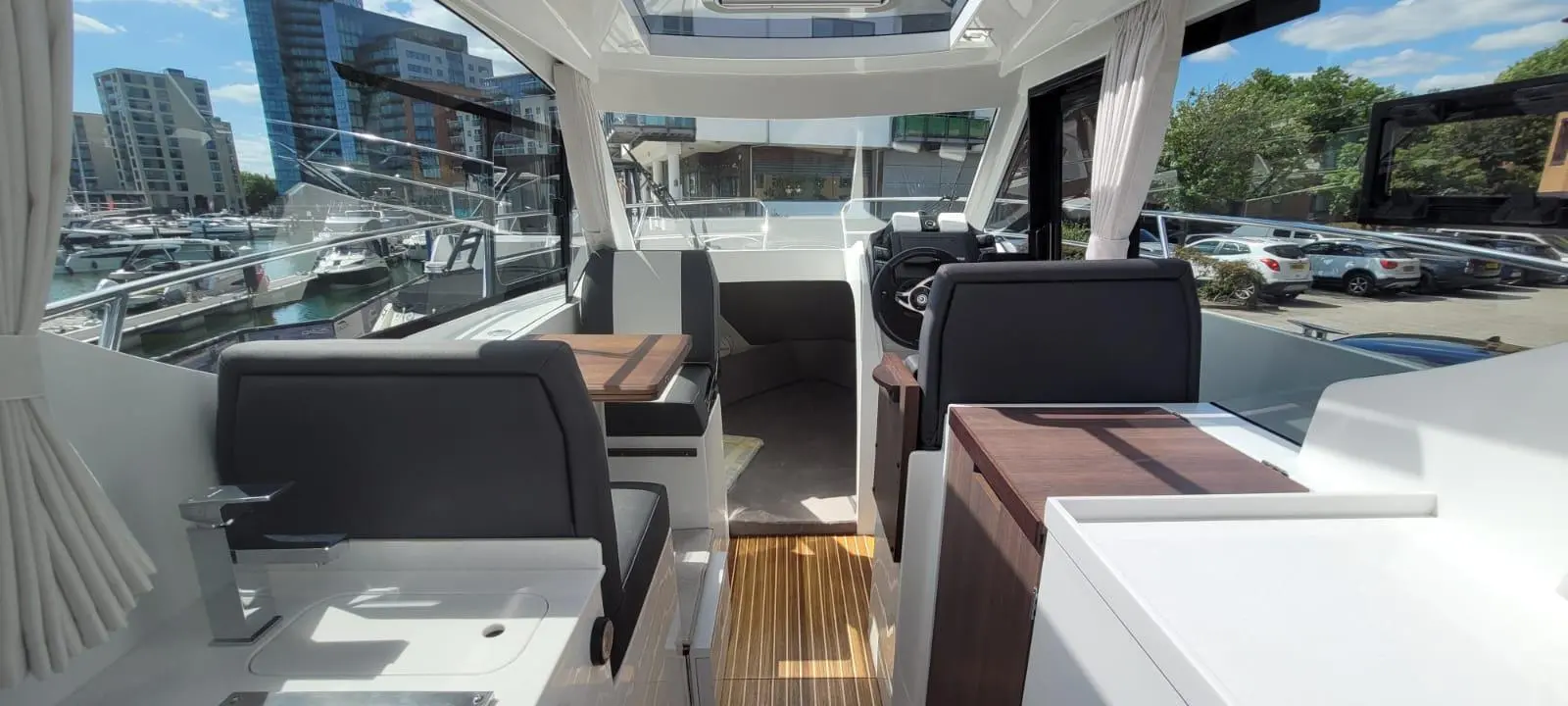 Interior view of a modern boat, featuring seating and a steering area, with a marina backdrop and blue skies.